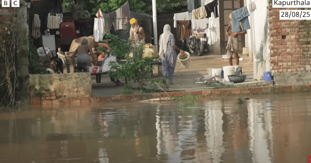 Punjab Floods 2025 "Flood-affected residents in Kapurthala, Punjab, standing outside their partially submerged homes with household items and clothes hanging to dry. (28 August 2025)"